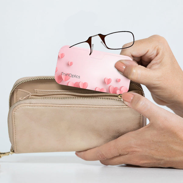 Brown ThinOptics Readers peeking out from a Pink Hearts Universal Pod being slipped into a clutch