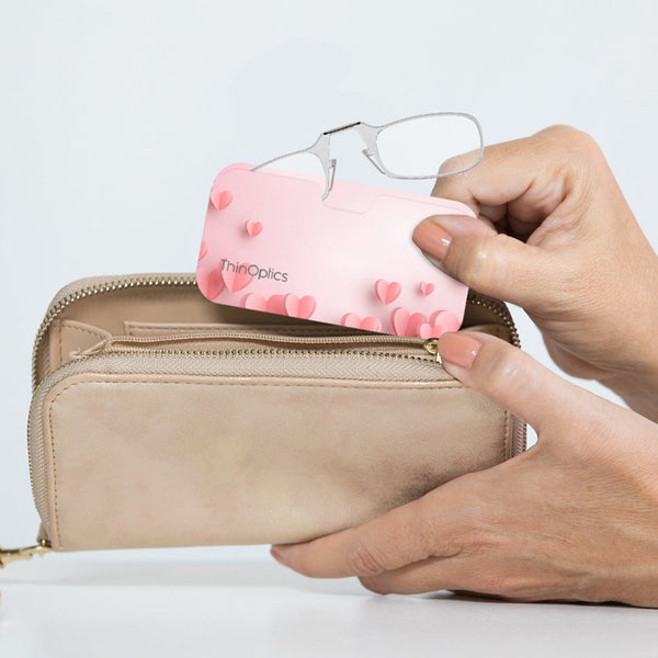 Clear ThinOptics Readers peeking out from a Pink Hearts Universal Pod being slipped into a clutch