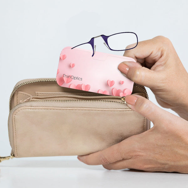 Purple ThinOptics Readers peeking out from a Pink Hearts Universal Pod being slipped into a clutch